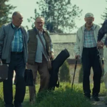 A group of elderly men in casual clothing carrying instruments in cases walk alongside a countryside road
