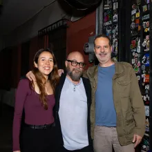 A young woman with long brown hair in a burgundy sweater poses arm in arm with two middle-aged men dressed casually in front of a local cinema