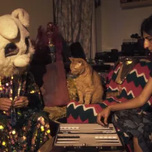 A figure in a colorful, sequined robe and bunny mask and a woman in a black shirt play backgammon. An orange cat sits between them, appearing to stare incredulously at the masked figure