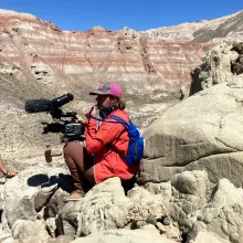 Woman in orange raincoat and pink cap holds a camera rig while drinking from straw connected to blue backpack. She sits on a boulder with a red striped cliff face behind her.