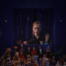 A blonde girl peers through spectacles into a space enclosed in purple curtains filled with various dolls