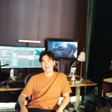 Young man in orange shirt sits in front of editing workstation with two large monitors