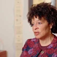 African American woman in her fifties, seated at a desk wearing a top featuring red and pink flower design. Picture frames are visible, but out of focus, in the background.