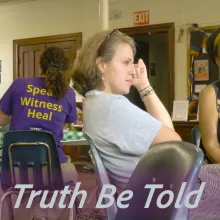 A man on the left in a black shirt at a table with a woman who is wearing a purple shirt with the words on the back: Speak, Witness, Heal.  A woman featured in the center of the photo is leaning back in her chair, legs crossed with her hand on her head looking off camera.  A black woman with a gold hair band is sitting next to the woman looking off camera.