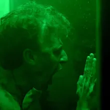 A white man presses against glass and screams, lit in bright green