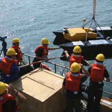 Eight men in orange vests and yellow hard hats on ship raise two containers out of the ocean