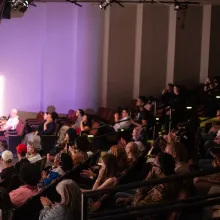 A packed theatre focuses on a stage which is off camera to the left. Purple light illuminates the audience