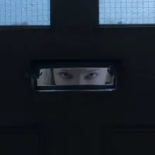 A blonde woman's blue eyes can be seen through a slot in an otherwise black surface with two windows visible at the top of the frame