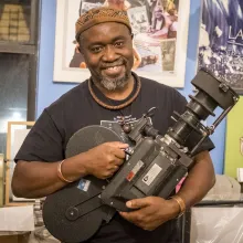 A smiling Black man in a brown hat, black T-shirt, and wooden and metal jewelry holds an old camera in front of awards and posters, one depicting Malcolm X