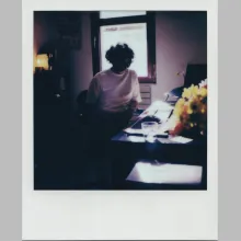 Polaroid of a man in a white T-shirt sitting at a desk which houses yellow flowers. The man is backlit by a window behind him and his face is obscured