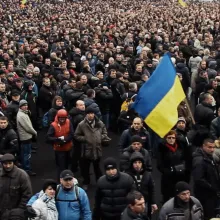 A massive crowd of people in winter clothes is gathered. One man towards the front waves a large Ukrainian flag