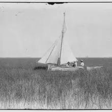 Black and white image of three men sitting in a white sailboat amidst marshland