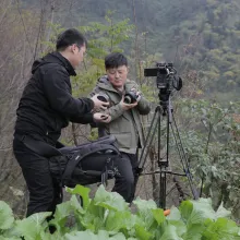 A man in a green jacket trades lenses with a man in all black. They stand behind a camera on a tripod amid forest