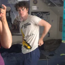 A young white man with short curly dark hair in an Andy Warhol white tee stares at a mirror in his bedroom as a white woman whose face is obscured by a camera takes a photo of them both