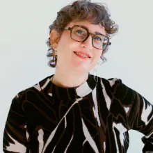 A woman with fair skin smiles at the camera. She has short, curly hair, green eyes, and is wearing a patterned shirt. 