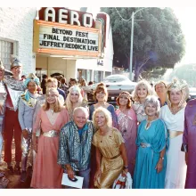 A row of 15 older white people in colorful robes stands in front of the AERO theatre, the marquee of which reads "BEYOND FEST"