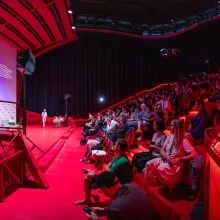 A large crowd in a theater lit in red looks at a big screen which displays a map of Czechia over a purple background