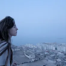 A woman in glasses with long brown hair looks over a city on the ocean at dusk