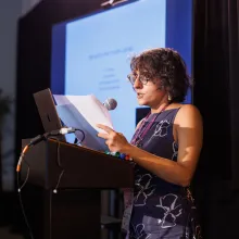 A woman (Sarvnik Kaur) with short, curly hair in a floral dress and glasses stands at a podium, speaking into a microphone. Behind her, out of focus, are a sign language interpreter and a large screen displaying text on a white background.