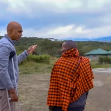 A tall man standing on crutches talks to a Maasai elder wearing a traditional shuka with small buildings behind them.