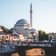 A place of worship in the city of Prizren, Kosovo, with a bridge in front of it from which a banner hangs reading "STORIES CONNECT US DOKUFEST." People flood the streets