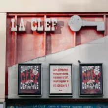 The front of a building, half red and half grey. At the top are the words "LA CLEF" and the shape of a key in grey. Three large posters are displayed on the building, reading "Ouverture La Clef Dēfinitive" and and "Un cinēma indēpendant, associatif, autogērē et solidaire!"