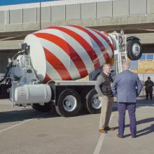 A concrete truck is seen parked on a concrete parking lot, with two white men, one in a suit, standing right by it