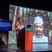 A man (Vinay Shukla) in a colorfully patterned shirt stands behind a podium and speaks into a microphone on stage. Behind him, a screen displays a poster of "An Insignificant Man" and an ASL interpreter, and a smaller screen provides captions of his words