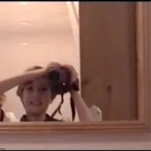 Pink tinted image of a smiling little girl with short hair filming herself in a mirror