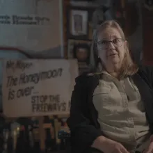 Image shows Kate Kelly, an older woman and resident of the brookland neighborhood who was a witness to the protests at the time. She is sitting being interviewed with a poster and signage from the protests blurred but still visible behind her. They say; "Mr. Mayor the honeymoon is over...stop the freeways!" and "White men's roads through black men's homes!"