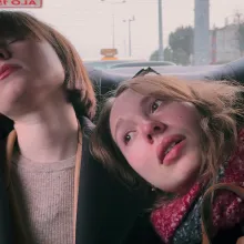 Two women in the backseat of a car, each with mid-length brown hair. The woman on the left, wearing a brown sweater under a black overcoat, sleeps, and the woman on the right, wearing a red scarf, rests her head on the other woman's shoulder and looks out the window