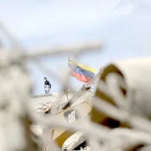  image - what can you physically see in this image (example below)?	A Venezuelan flag flies amid rubble and heavy machinery, with a person wearing a face mask standing nearby. Out-of-focus debris frames the foreground.