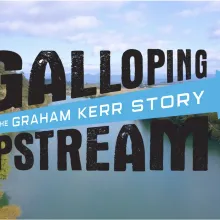 A cinematic aerial view of a winding river surrounded by lush green forests and open farmland under a blue, cloud-filled sky. Bold, distressed black lettering reads “GALLOPING UPSTREAM,” crossed by a bright turquoise arrow containing the white subtitle “THE GRAHAM KERR STORY.” The design feels adventurous, energetic, and inspirational, suggesting a journey of perseverance and discovery.
