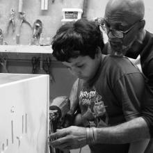 :A  bald Black man with glasses teaches a young boy with black hair how to engrave a casket with a drill.