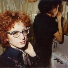 A woman wearing black adjusts her eye makeup in the mirror, while a red-headed woman wearing glasses looks at the camera in the foreground. From ‘All the Beauty and the Bloodshed.’ Photo courtesy of Neon. 