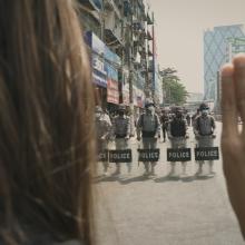A person holds up three fingers standing in front of a line of police in riot gear. From ‘Myanmar Diaries.’ Photo courtesy of Double Exposure Film Festival. 