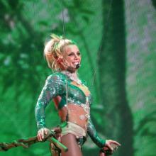 Pop star Britney Spears, subject the recnt documentary 'Framing Britney Spears," is pictured performing on stage, wearing an emerald green sequin outfit, singing into a wireless headset, clutching a brown railing, against a mesh, emerald green patterned backdrop.