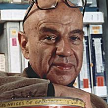 A middle-aged Bud Greenspan posing in front of film tapes. He is bald and is wearing black framed glasses on his head.