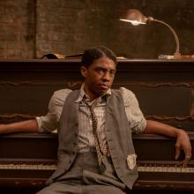 Actor Chadwick Boseman, a Black man wearing a striped shirt, patterned tie, grey vest and pants, sits at a piano, his back to the keyboard
