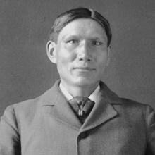 A black and white portrait of an Indigenous man with short hair wearing a suit. 