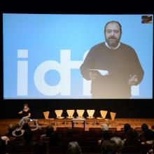 IDFA Artistic Dierctor Orwa Nyrabia, shown delivering a keynote address on a large screen at the 2018 Getting Real conference, is a Syrian man with a beard, wearing a dark sweater. Photo courtesy of AMPAS