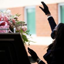 The photo depicts a Black woman standing over an open casket, her right arm raised in praise; she wears a protective face mask, as well as a dark suit.