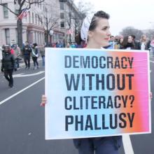 A woman is particpating in a demonstration on a city street; she carries a sign that reads "Democracy without cliteracy? Phallusy."