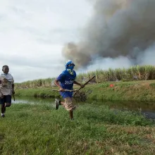 Two Black teenage boys run across a green field holding sticks. Smoke bellows behind them. Still from Patrick Bresnan's 'The Rabbit Hunt'. Courtesy of Argo