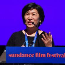 Jean Tsien is a female Asian American filmmaker seen here receiving the Art of Editing Mentorship Award at Sundance, 2018. She is speaking into a mic wearing a black sweater. Photo courtesy of Jean Tsien.
