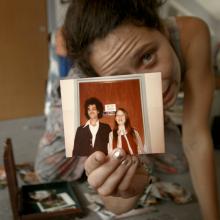 A white woman with dark hair crouched over holding up a polaroid of a man and a woman. A still from Nira Burstein's 'Charm Circle'. Courtesy of Big Sky Documentary Film Festival. 