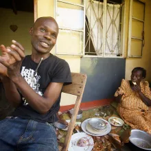 David Kato, a Black man with a bald head sits in front of a yellow house. His mother, Lydia Mulumba Nalongo, an older Black woman sits behind him eating. A still from 'Call Me Kutchu,' courtesy of the filmmakers.