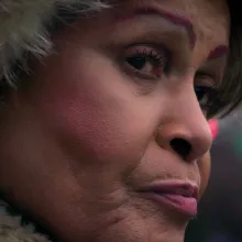 Close-up of Black trans woman, Gloria Allen, standing at a bus stop, wearing long oval silver earrings. Film still from Luchina Fisher's 'Mama Gloria'.
