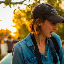 Headshot of a woman in her late 20s wearing a baseball cap over short brown hair. She has a camera hanging around her neck and it is golden hour behind her through the trees. 