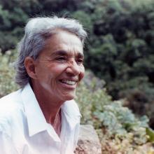 Chavela Vargas is a queer Latin American singer with grey hair. She is wearing a white shirt and is smiling. She is surrounded by trees and shrubs. Image from Catherine Gund and Daresha Kyi’s ‘Chavela.’ Courtesy of Alicia Elena Pérez Duarte, Aubin Pictures.